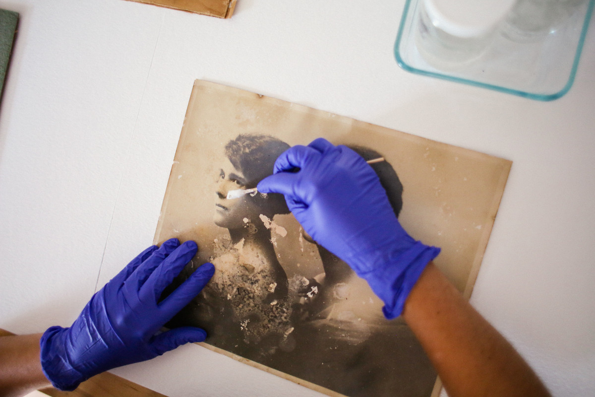 This image shows two hands wearing blue nitrile gloves, working on a silver gelatin print, depicting two woman in bust. The right hand is holding a Q tip used to clean the photograph. The print shows stains and image loss caused by mould damage.