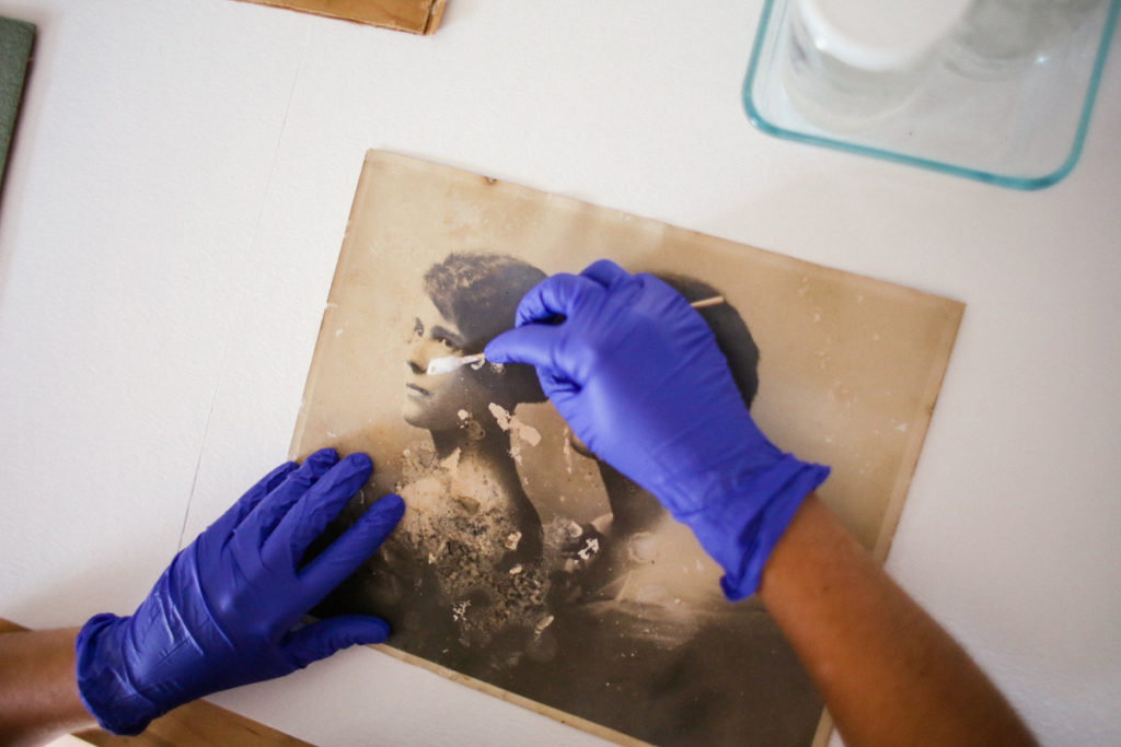Cette image montre deux mains portant des gants en nitrile bleu, travaillant sur une épreuve en gélatine argentique, représentant deux femmes en buste. La main droite tient un embout Q utilisé pour nettoyer la photographie. Le tirage montre des taches et des pertes d'image causées par des moisissures.