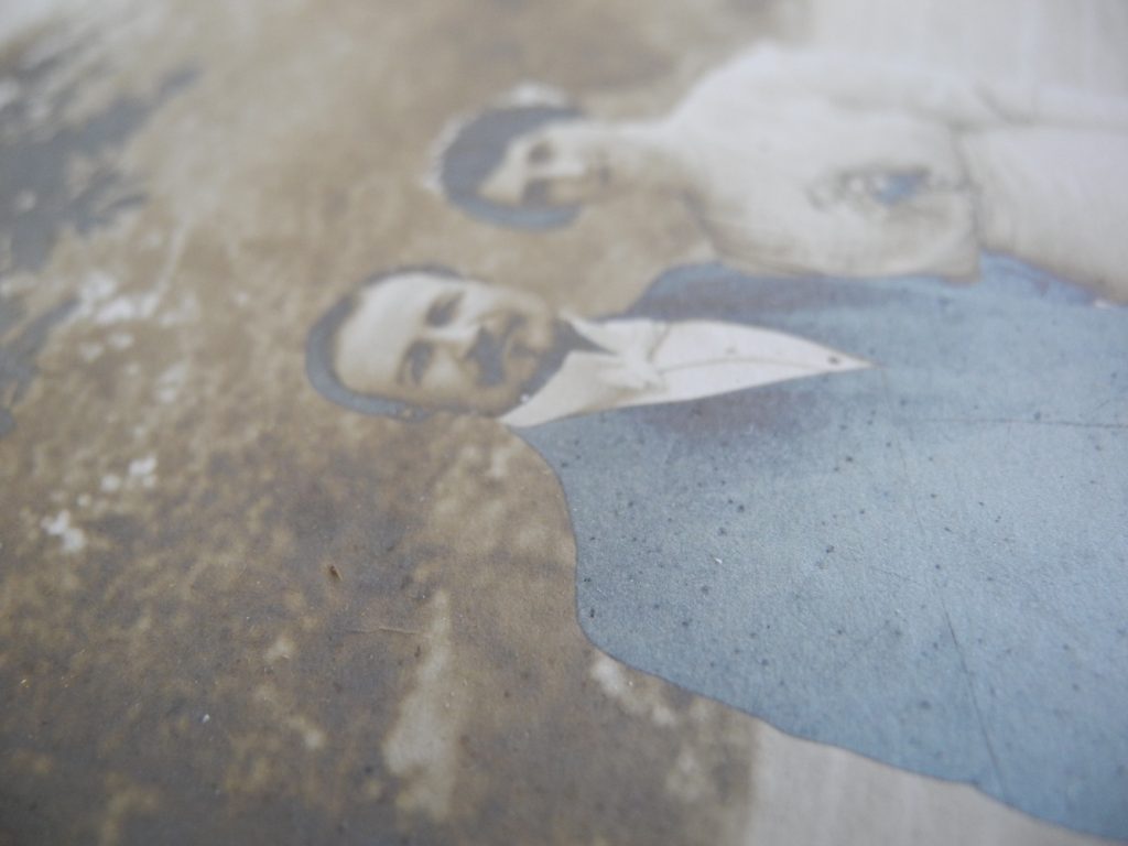 Détail d'une photographie gélatino-argentique prise en lumière rasante. La photographie représente la un couple de marié, l’homme portea un costume et la femme porte une robe blanche, l’image est recadrée au niveau de leur taille. Le tirage présente un fort affaiblissement, jaunissement et miroir d’argent.