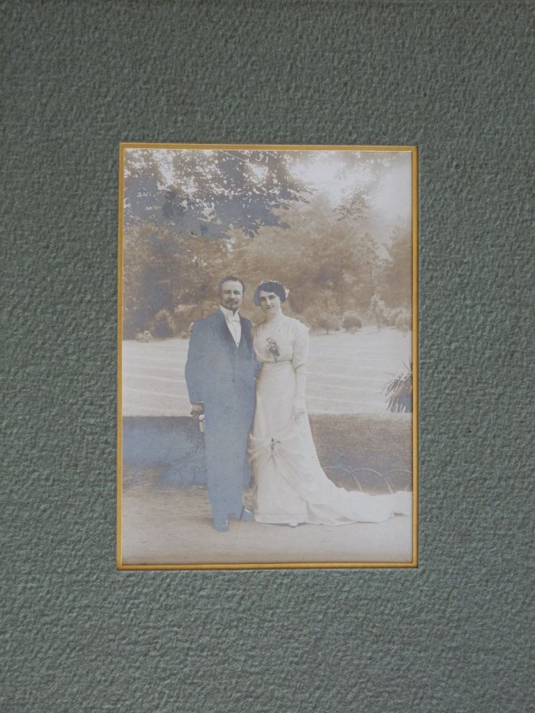 A silver gelatin photograph mounted with a forest green texture window mat, with a golden rim on the opening. The photograph depicts a couple’s wedding picture of a man wearing a suit and a woman wearing a white dress, they are standing in front of a lawn. The print has extensive fading, yellowing and silver mirroring.