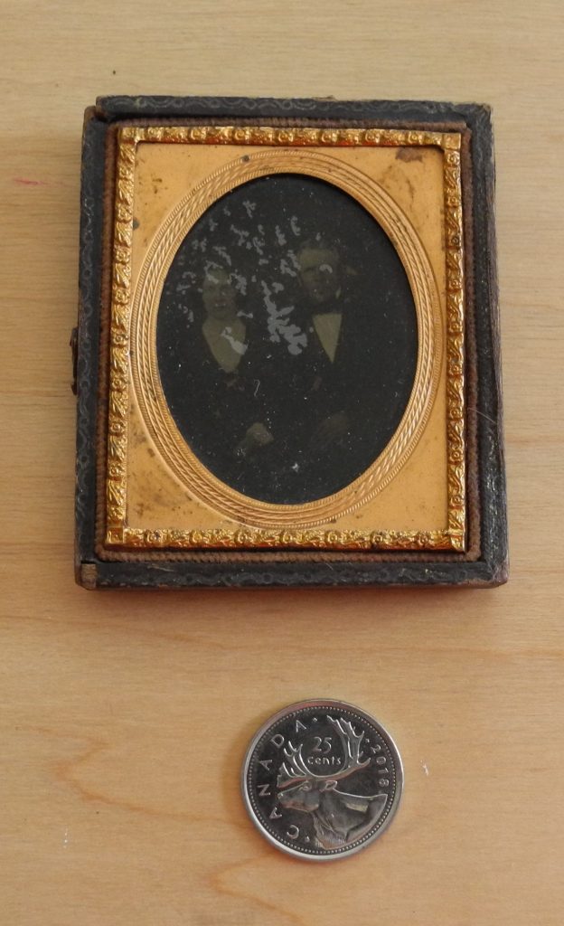 Vue en plongée d'un ambrotype, représentant le portrait d'un homme et d'une femme jusqu'aux genoux, monté dans un passe-partout et un preserver en laiton doré, dans un demi-écrin de style américain. Une pièce canadienne de 25 sous est placée le long du bord inférieur de la photographie.