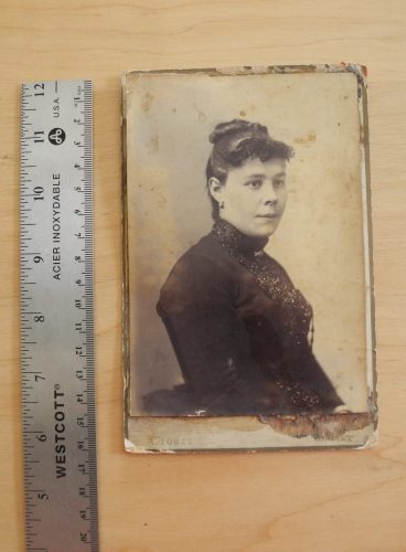 Overhead image of a monochrome photograph, depicting a bust-length portrait of a woman, mounted on a beige cabinet card. A metallic ruler is placed along the left side of the photograph.